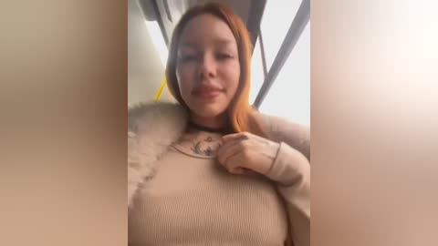 A video of a smiling young woman with fair skin and shoulder-length, reddish-brown hair, wearing a beige ribbed sweater. She is seated indoors, possibly on public transportation, with a blurred background.