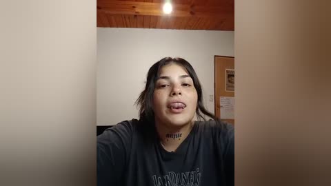 Media: Video of a young woman with long dark hair, sticking out her tongue, wearing a black shirt with \"Hollywood\" in white letters. She has a small tattoo on her neck. Background shows a wooden ceiling and wall with a framed picture.