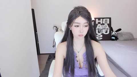 Media: Video of a slender, light-skinned Asian woman with long, straight black hair and a purple halter top, sitting on a white chair in a minimalist bedroom with a bed and wall art.