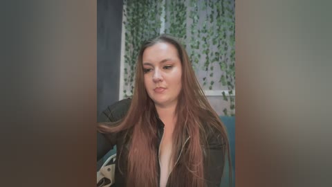 Video of a Caucasian woman with long, straight, reddish-brown hair, wearing a black jacket, sitting indoors. The background features a green ivy wall and a blue couch.