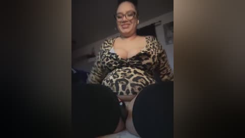 Video of a plus-sized woman with light skin and dark hair, wearing leopard print lingerie and black thigh-high stockings, sitting on a bed in a dimly lit room.