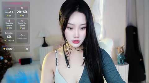 Media: Video of a young East Asian woman with long black hair and pale skin, wearing a gray top and a black choker, standing in a softly lit bedroom.