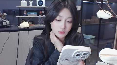 Video of an Asian woman with long black hair, wearing a black top, using a white iPhone, sitting in a modern office with white shelves, a desk lamp, and a black wall.