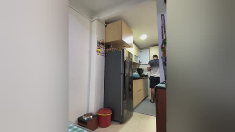 Video of a small, compact kitchen with light wood cabinets, a black refrigerator, a red bucket, and a person in the background.