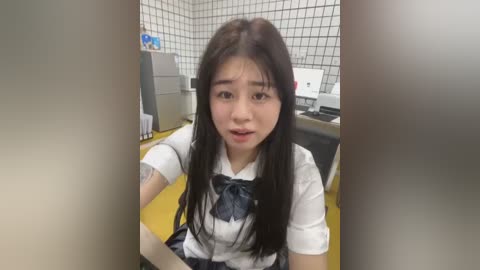 Media: Video of an Asian woman with long black hair and a white blouse, standing in a tiled room with a yellow floor, near a computer monitor and a refrigerator.