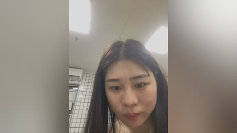 Media: Video of an Asian woman with long black hair and fair skin, looking serious, in a bathroom with white tiles and a vent.