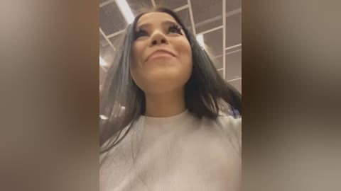 Video of a young woman with long, straight black hair, light brown skin, and a confident expression. She wears a white top in an indoor setting with a grid ceiling.