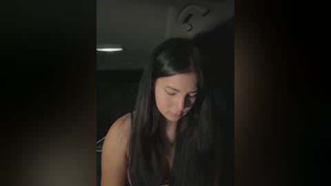 Media: A video shows a young woman with long, straight black hair, wearing a black top, standing in a dimly lit room, looking down with a thoughtful expression.