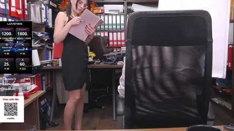 Media: Video of a woman in a black dress, holding a pink binder, standing in a cluttered office with shelves full of books, a desk, and a black chair.