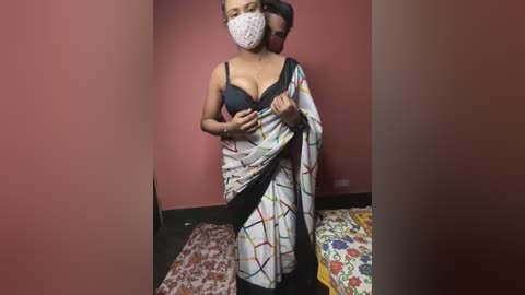 Media: Video of a South Asian woman in a black bra, white polka-dot face mask, and colorful sarong, standing in a small, red-walled room with a floral-patterned bedspread.