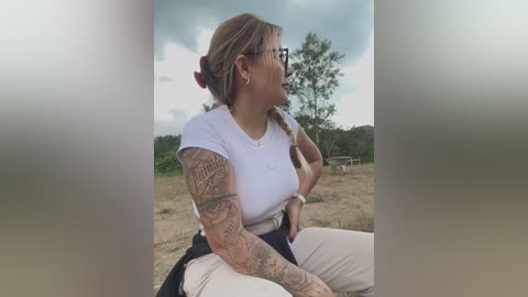 A video of a tattooed woman with blonde hair, wearing glasses, a white t-shirt, and beige pants, sitting outdoors with a cloud-filled sky and green trees in the background.