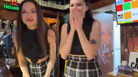 Media: Video of two young women with fair skin, straight dark hair, wearing black crop tops and plaid skirts, standing in a cozy bar with Heineken sign, colorful posters, and a TV screen.