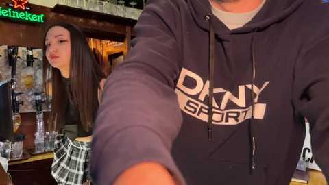 Media: A video of a young woman with long brown hair in a black hoodie, standing behind a bar counter. The background features a dimly lit bar setting with a green neon sign.