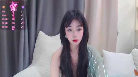 Media: A video of an East Asian woman with long black hair, wearing red lipstick, sitting on a bed with white sheets and pillows.