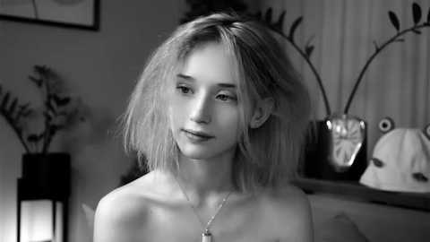 Media: Black-and-white video of a young, slender Caucasian woman with shoulder-length, tousled hair and a delicate facial structure, wearing a necklace. She is indoors, surrounded by potted plants and decorative objects on a wooden shelf.