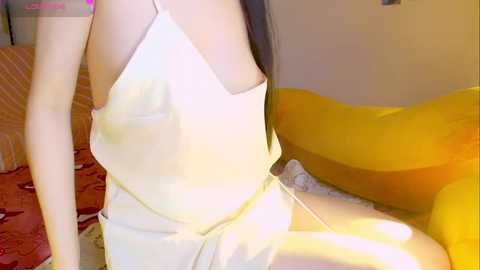 Video of a slender East Asian woman with long black hair, wearing a white, spaghetti-strapped nightgown, sitting on a yellow beanbag chair in a dimly-lit, cozy room.