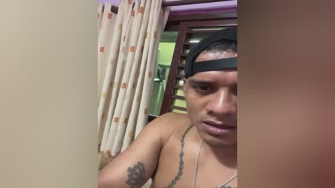 Media: A video of a young, shirtless Latino man with short hair, wearing a black baseball cap, and a necklace, standing indoors. He has tattoos on his arm. The background features light-colored curtains and a wooden door.