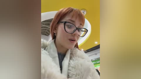 Video of a young woman with short, reddish-brown hair and glasses, wearing a white fur-trimmed coat, in a brightly lit, modern indoor setting with yellow and white walls.