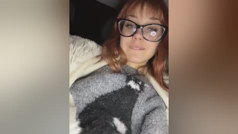 Video of a Caucasian woman with shoulder-length auburn hair, wearing black-rimmed glasses and a gray argyle sweater, lying on a white blanket in a dimly lit room.