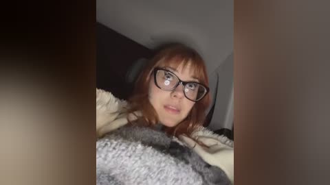 Video of a young Caucasian woman with short red hair, wearing glasses and a gray sweater, lying on a bed with beige sheets. The background is dimly lit, suggesting a bedroom setting.