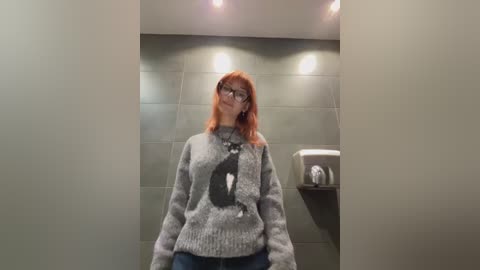 A video of a middle-aged woman with shoulder-length red hair, wearing glasses and a gray, patterned sweater with a large, black, cartoonish wolf graphic. She stands in a modern, gray-tiled bathroom with recessed lights and a paper towel dispenser on the right.