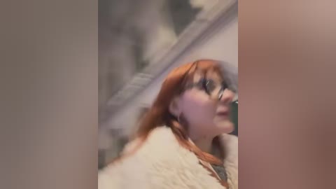 A blurry video of a woman with long red hair and glasses, wearing a white sweater, standing in a dimly lit room with indistinct furniture and lighting.