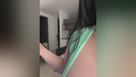 A video shows a woman with long, straight black hair, wearing a green tank top, standing in a modern, dimly-lit living room with beige carpet and white walls.