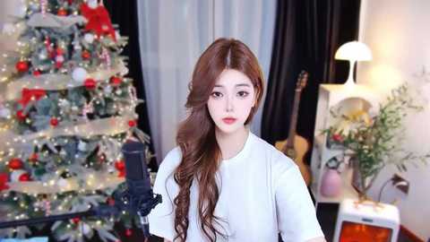 Video of a young Asian woman with long, wavy brown hair, wearing a white blouse, standing in a festive room with a decorated Christmas tree, a guitar, and a lamp.