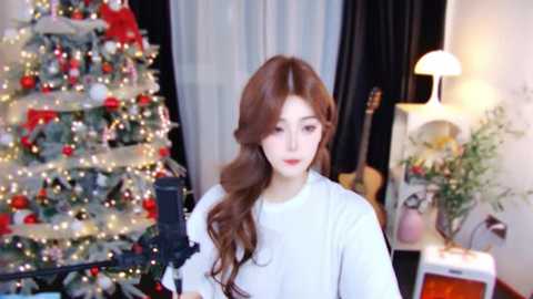 Video of a young woman with long, wavy auburn hair, wearing a white sweater, standing in a cozy room with a lit Christmas tree and a warm lamp.