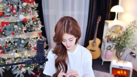 Media: Video of a young woman with long, wavy brown hair singing into a microphone in a cozy, festive room with a decorated Christmas tree, guitar, and potted plants.