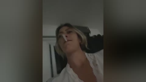 Media: Video of a blonde woman with light skin, wearing a white lace top, smoking a cigarette indoors, dim lighting, blurred background, intimate and moody atmosphere.