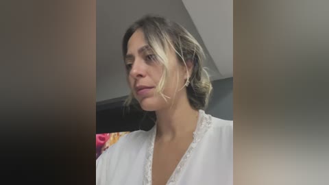 Media: Video of a woman with fair skin, brown hair in a messy bun, wearing a white lace-trimmed blouse, looking contemplative, blurred background with colorful clothes.