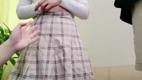 Video of a person in a white long-sleeved shirt and plaid skirt, adjusting the waistband with hands. Background features a green plant and beige wall.