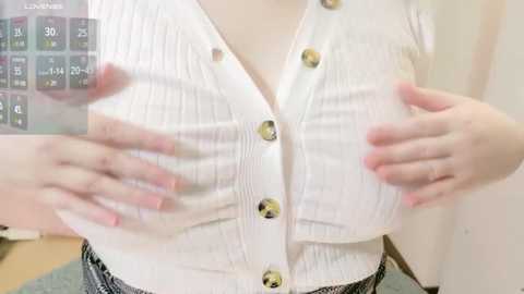 Media: Video of a fair-skinned woman with medium breasts, wearing a white, button-up cardigan with visible nipples, hands cupping her chest. Background shows a digital clock and a blurred room.