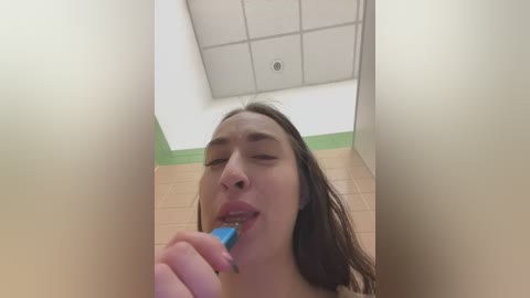 Video of a young woman with long brown hair, fair skin, and a neutral expression, brushing her teeth in a public restroom with beige tiles and a fluorescent ceiling.