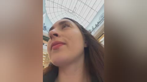 A video of a woman with light skin, long brown hair, and a confident smile, standing in a modern, glass-roofed shopping mall. The image is taken from a low angle, emphasizing her upward gaze.