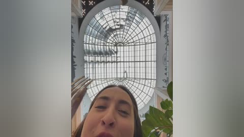 Video of a young woman with long brown hair, standing under an ornate, circular glass dome ceiling with intricate metalwork, in a grand, possibly European-style building with white columns.