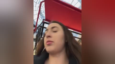 Video of a young woman with long brown hair, fair skin, and closed eyes, standing under a red canopy in an indoor space with a glass ceiling and metal structures.