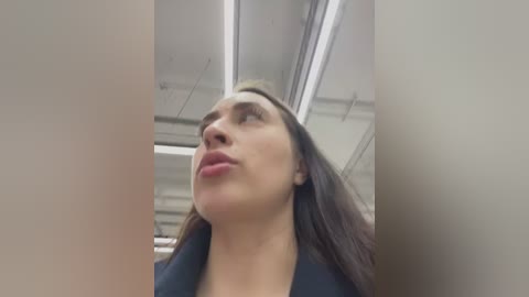 Video of a woman with long, straight brown hair, wearing a black top, looking upward in an office with fluorescent lights and white walls.