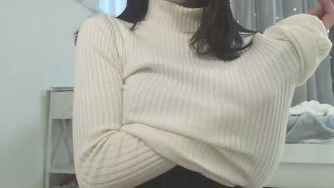 Video of a woman in a cream ribbed turtleneck sweater, with long dark hair, standing indoors. The background features a white wall, a bed with gray sheets, and a window with sheer curtains.