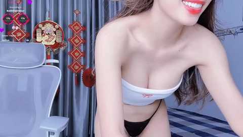 Media: A video of a smiling Asian woman with fair skin, wearing a white bandeau top and black panties, standing in a room with gray curtains and colorful Asian decorative items.