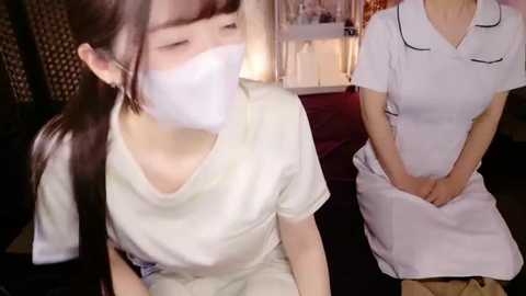 Video of two Asian women in white medical scrubs and masks, seated on a couch in a dimly lit room, hands clasped.