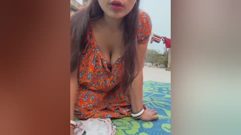 Media: Video of a woman with medium brown skin, long dark hair, wearing an orange floral dress with a low neckline, kneeling on a green and blue patterned towel, with a man in the background.