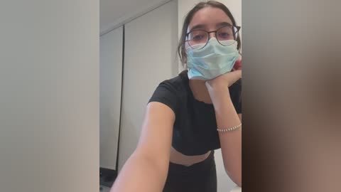 Media: Video of a young woman with light skin, wearing glasses, a black shirt, and a blue face mask, standing in front of a white closet, her arms extended towards the camera.