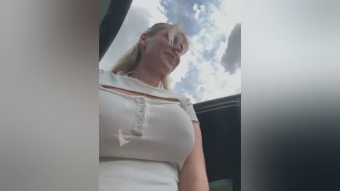 Media: A video of a smiling, blonde woman in a white ribbed top, viewed from below. The background features a partially open car door and a cloudy sky.