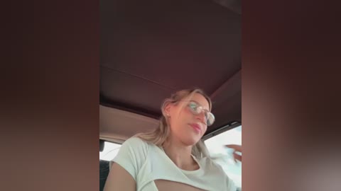 Media: A video of a woman with light skin and blonde hair wearing glasses and a white shirt, captured from a car window, showing a blurred, dark-tinted interior.