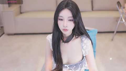 Video of a young East Asian woman with long black hair, wearing a low-cut white dress with floral patterns, seated indoors on a light-colored floor, surrounded by modern furniture.