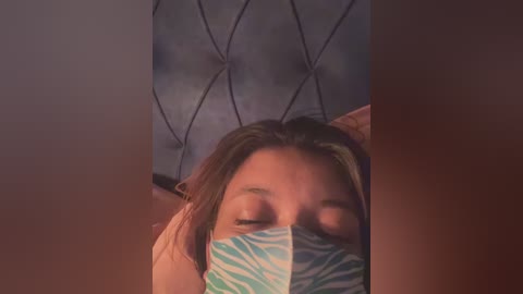 Media: Video of a young woman with light skin, wearing a blue and white face mask, lying on a diamond-patterned quilted bed, eyes closed, appearing calm and relaxed.