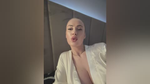 Video of a fair-skinned woman with blonde hair, wearing a white silk robe, making a puckered lip pout. Background features a gray upholstered headboard.