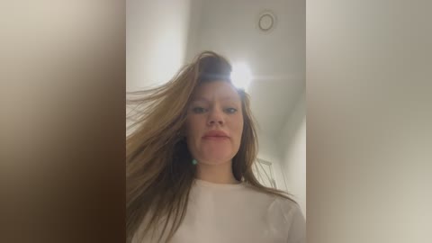 Video of a Caucasian woman with light skin and long brown hair, wearing a white shirt, indoors, blurred background, slightly out of focus, looking serious.